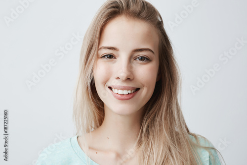 Pleasant-looking woman with dark eyes, beautiful lips and pure skin looking with smile at camera rejoicing her vacation, relaxing. Positive female with long blonde hair posing against gray wall