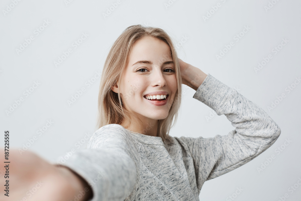 Attractive dark-eyed blonde female dressed casually having delightful look smiling broadly. Beautiful woman having cheerful expression while posing against gray background, stretching arm to camera. Attractive dark-eyed blonde female dressed casually having delightful look smiling broadly. Beautiful woman having cheerful expression while posing against gray background, stretching arm to camera.