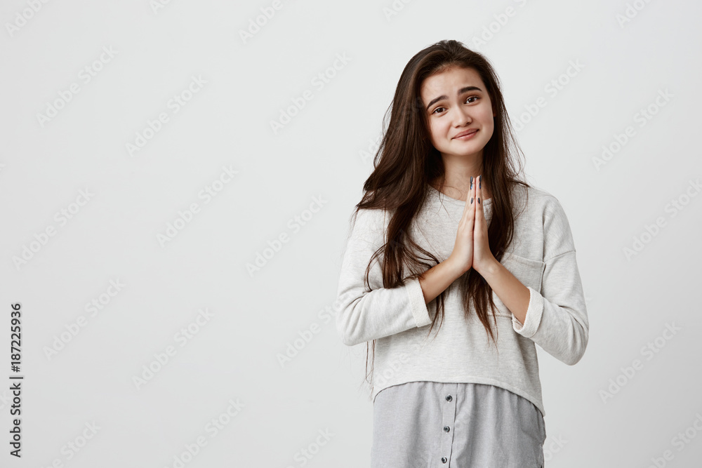 Beautiful girl with dark eyes and healthy skin, smiling, having hopeful expression keeping hands together asking for something. Natural brunette female anticipating for something important in life Beautiful girl with dark eyes and healthy skin, smiling, having hopeful expression keeping hands together asking for something. Natural brunette female anticipating for something important in life