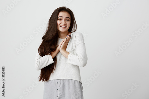 Beautiful young female with dark eyes, healthy skin having surprised and excited expression keeping hands together smiling broadly. Happy glad girl with long dark hair expressing positive emotions