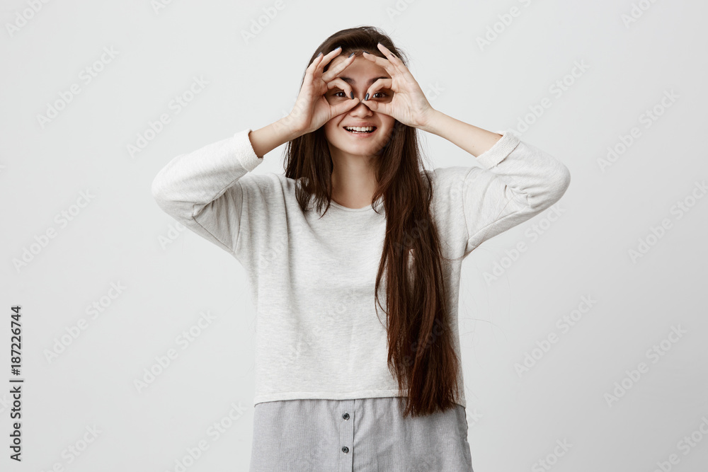 Cheerful excited teenage female with dark long straight hair showing Ok gestures with both hands, pretending to wear spectacles, and smiling broadly, enjoying her carefree happy life Cheerful excited teenage female with dark long straight hair showing Ok gestures with both hands, pretending to wear spectacles, and smiling broadly, enjoying her carefree happy life