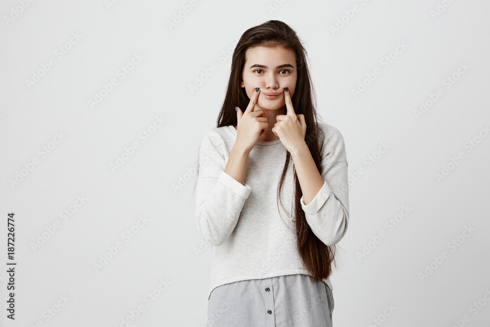 Headshot of young upset brunette female teenage making fake smile with her fingers stretching the corners of her mouth. Portrait of school girl trying to stay positive after failing exams at school Headshot of young upset brunette female teenage making fake smile with her fingers stretching the corners of her mouth. Portrait of school girl trying to stay positive after failing exams at school