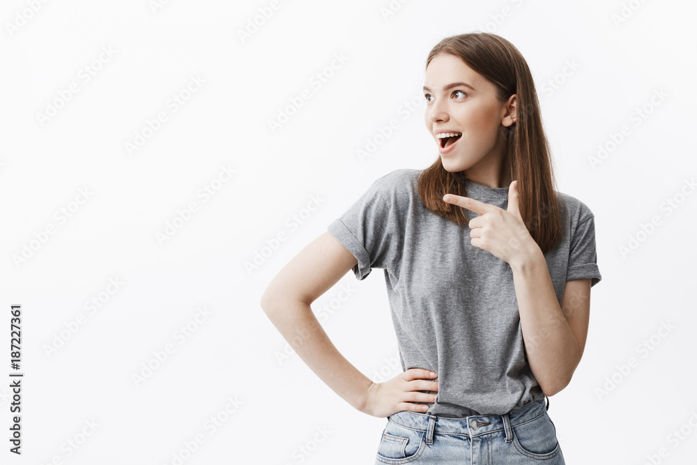 Copy space. Good-looking cinfident young european student with dark hair in casual grey clothes looking aside with opened mouth and excited expression, pointing with finger on free space on white Copy space. Good-looking cinfident young european student with dark hair in casual grey clothes looking aside with opened mouth and excited expression, pointing with finger on free space on white
