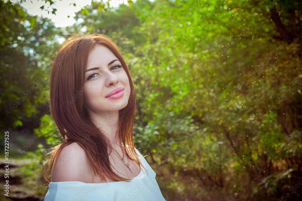 Portrait of beautiful young smiling woman with long red hair, wearing elegant white top in a forest with green trees on a sunny summer day Portrait of beautiful young smiling woman with long red hair, wearing elegant white top in a forest with green trees on a sunny summer day
