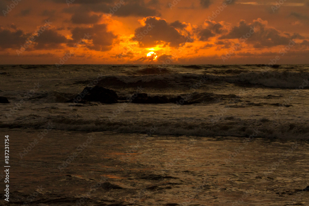 Obraz premium Waves crash ashore as the Southern Pacific sun sets in the background, turning the sky to liquid orange fire that is reflected in the water.