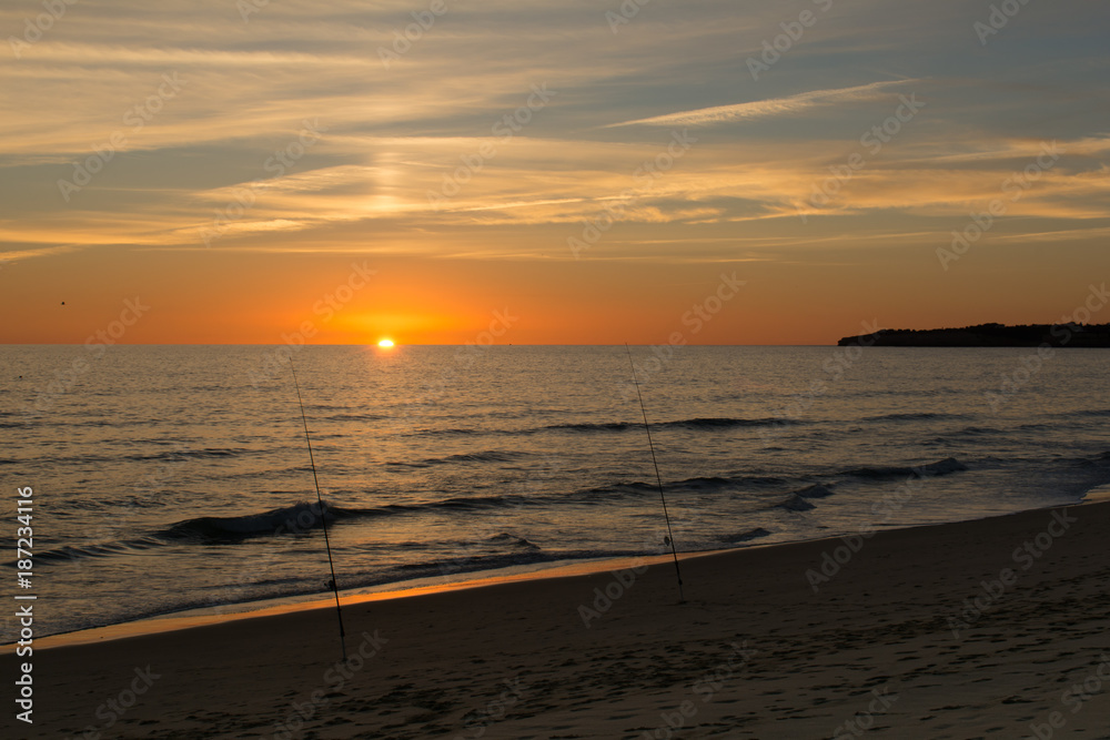 Sun dipping into the ocean with twa fishing rods in the foreground