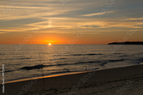 Sun dipping into the ocean with twa fishing rods in the foreground