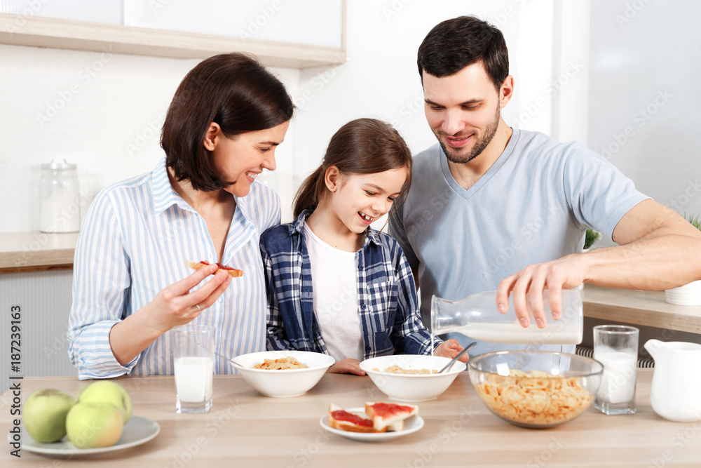 Happy young parents and their lovely daughter sit together at kitchen table, eat flakes, have healthy breakfast, enjoy good morning, have friendly relationship. Family and eatting concept. Happy young parents and their lovely daughter sit together at kitchen table, eat flakes, have healthy breakfast, enjoy good morning, have friendly relationship. Family and eatting concept.