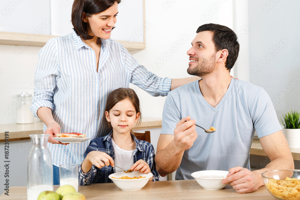 Three family members have delicious healthy breakfast at kitchen, eat cornflakes with milk, enjoy togetherness and domestic atmosphere. Husband looks with love at wife, satisfied with tasty dish.