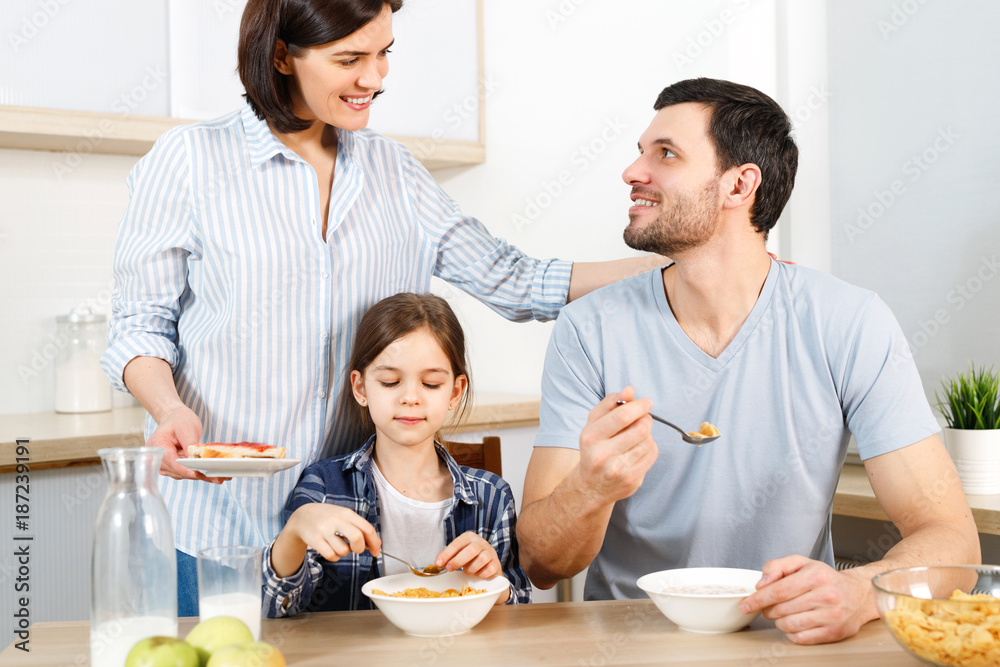 Three family members have delicious healthy breakfast at kitchen, eat cornflakes with milk, enjoy togetherness and domestic atmosphere. Husband looks with love at wife, satisfied with tasty dish. Three family members have delicious healthy breakfast at kitchen, eat cornflakes with milk, enjoy togetherness and domestic atmosphere. Husband looks with love at wife, satisfied with tasty dish.