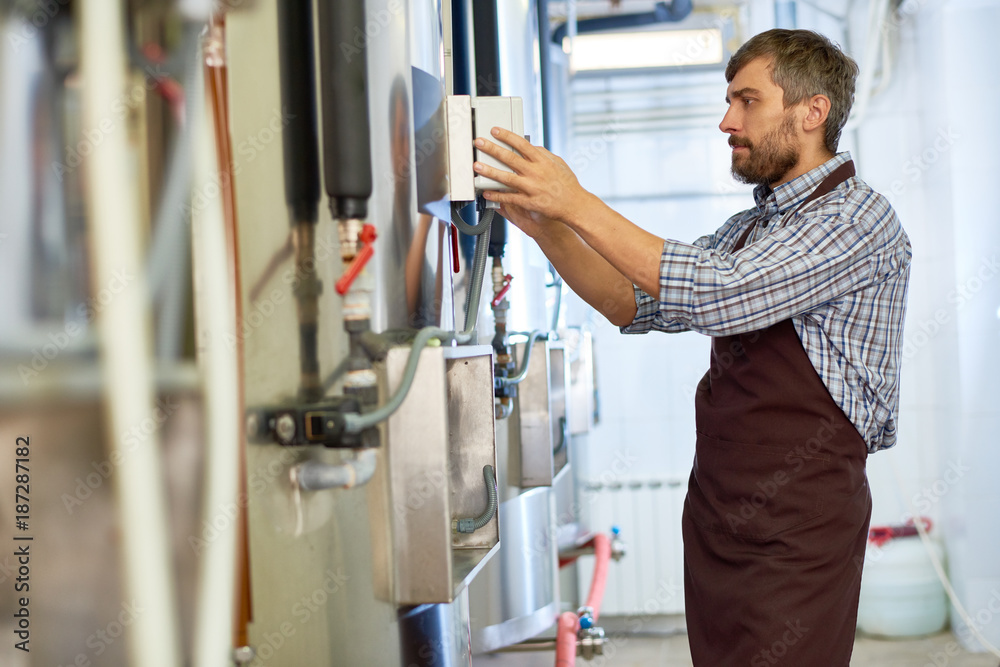 Profile view of confident middle-aged engineer wearing checked shirt and apron adjusting equipment while standing at modern brewery, portrait shot