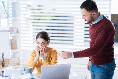 Effective treatment. Tired ill woman sitting in front of a laptop and holding a napkin near the nose while her friendly attentive supporting colleague giving her effective nasal drops