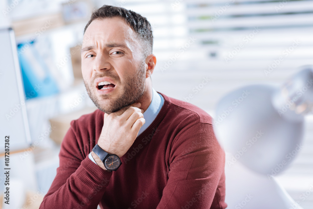 Naklejka premium Sore throat. Young bearded worker sitting in his light big office and touching his neck while having an awful sore throat