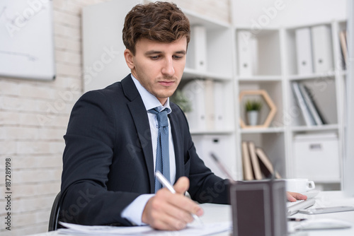 Portrait of young handsome businessman writing notes while using computer at desk while working in modern in office