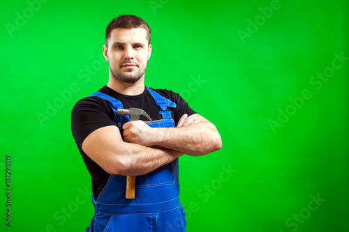 A dark-haired male construction worker in a black T-shirt and blue construction overalls smiling, posing and  holding wood hammer  on a green isolated background