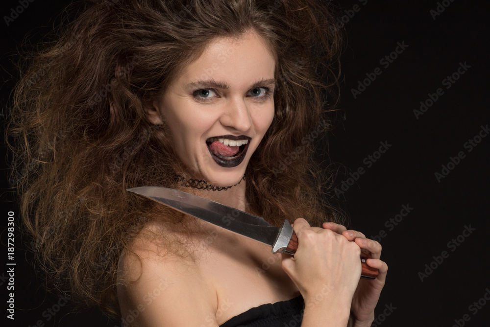 Obraz premium Beautiful girl with makeup as a witch smiles and holds a knife on a black background for the holiday of Halloween