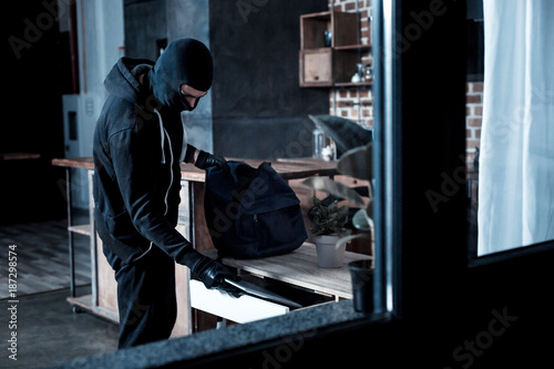 Professional burglar. Violent professional masked burglar wearing a black uniform and gloves and stealing a laptop from the table and holding a bag photo