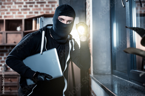 Stealing laptop. Angry professional masked criminal wearing black gloves and holding a torch while stealing a laptop