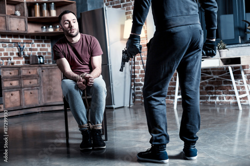 Killer. Shocked alarmed bearded man sitting with his hands tied while a burglar standing near him and holding a gun