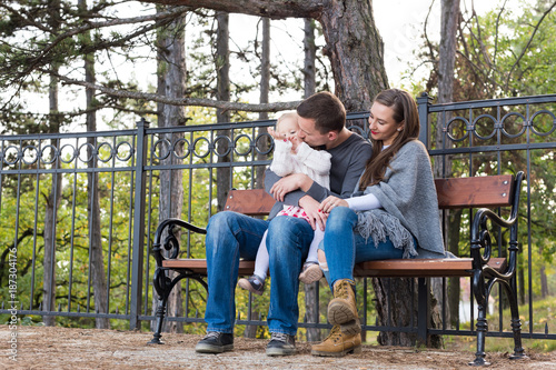 Happy family of three sitting on a bench in a park enjoying their time together. Father giving a kiss to his toddler daughter, mother smiling.