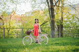 Young smiling girl standing near bicycle with flowers in park. Bikes bicycle girl. Teenager wearing pink polka dots dress looking dreamily. Romantic style. Happy and free lady.