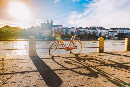 city bicycle with basket on the steering wheel of red color on the quay near the river Rhine in Switzerland against the backdrop of old city and authentic houses and cathedral on sunny day in winter