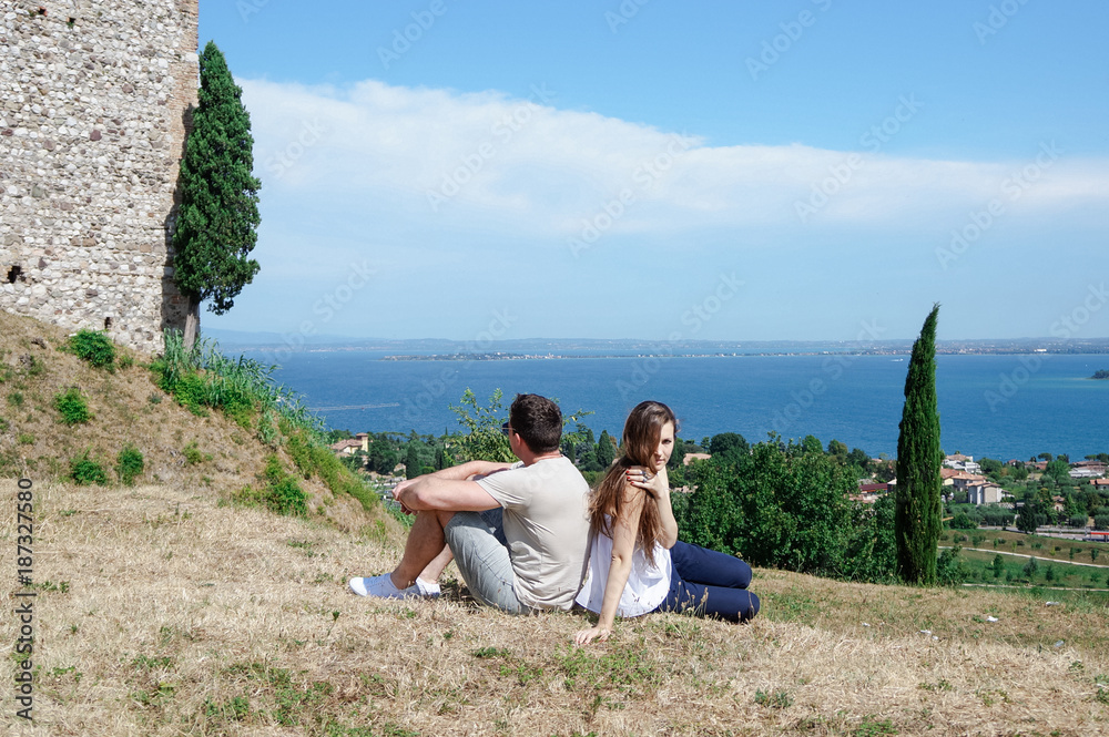 Handsome man and a young woman sitting next to him on the dried grass near an old castle on a hill overlooking the sea and the blue sky