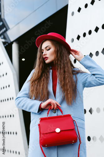 Outdoor waist up portrait of young beautiful fashionable woman posing in street. Model wearing light blue coat, red hat, holding trapeze handbag. Female fashion concept