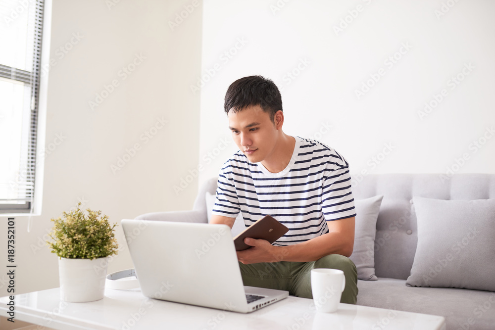 Handsome serious man, looking at laptop screen and writing something in his notebook, sitting on couch Handsome serious man, looking at laptop screen and writing something in his notebook, sitting on couch