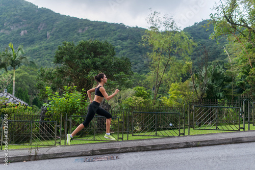 Young fit woman doing cardio exercise, listening to music, running outdoors with green mountain landscape in the background © undrey