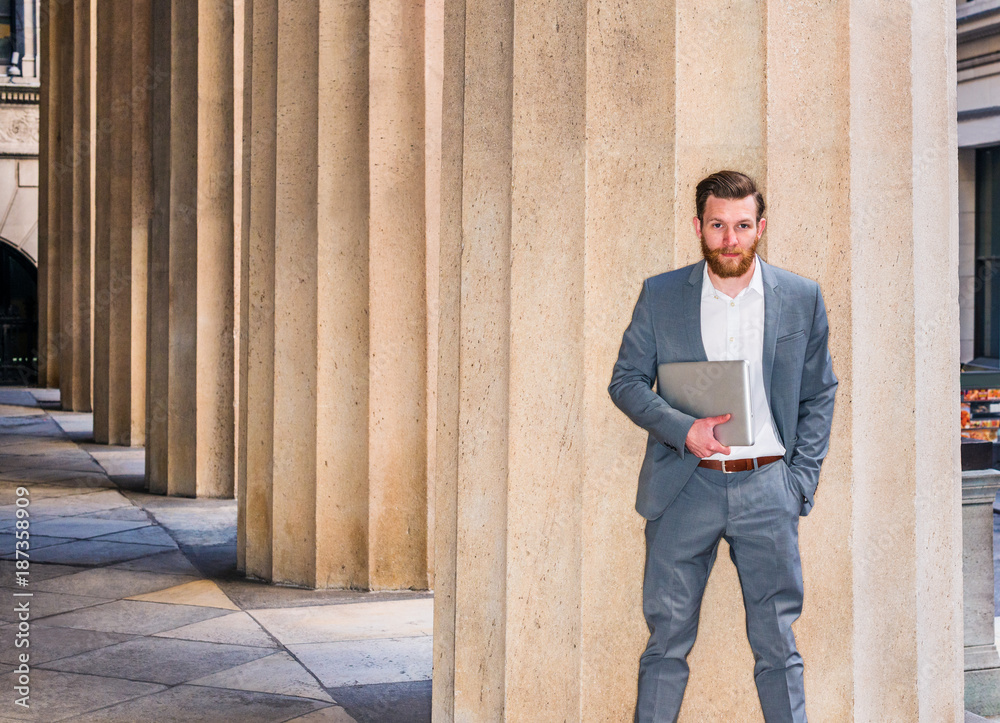 American Businessman with beard, mustache working in New York, wearing cadet blue suit, white undershirt, carrying laptop computer, standing against column outside office building, looking at you..