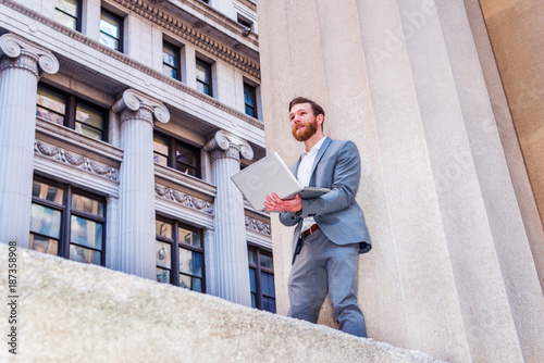 American Businessman with beard, mustache working outside in New York, wearing cadet blue suit, white undershirt, standing against column on street with vintage buildings, working on laptop computer