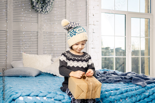 little girl the child sitting in pajamas and hat on the bed with garland of light bulbs with gifts boxes wrapped in a non-colored paper decorated with cones on blue knitted coverlet.Christmas concept photo