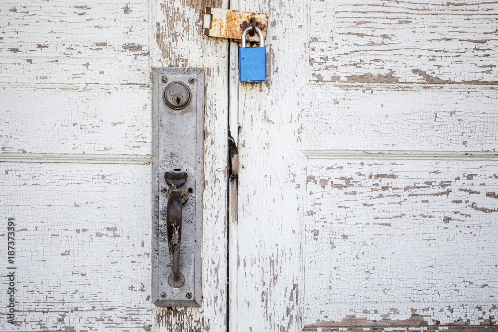 Padlock on a rusty hinge on peeling white wooden double doors above a metal door handle Padlock on a rusty hinge on peeling white wooden double doors above a metal door handle