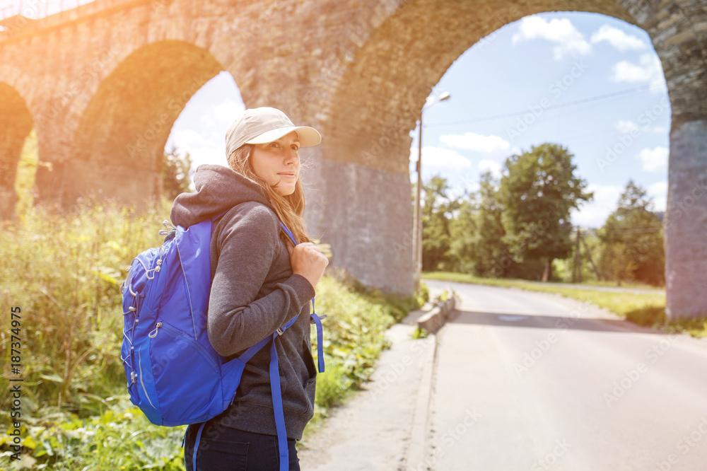 Young hitchhiker woman with backpack standing near old viaduct. Tourist backpacker girl in scenery country side