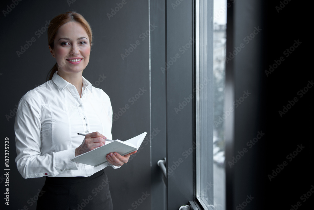 Portrait of cheerful businesswoman writing in appointment book while looking at window. Occupation concept Portrait of cheerful businesswoman writing in appointment book while looking at window. Occupation concept