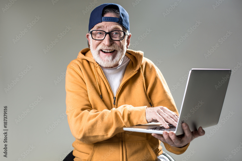 Waist up portrait of trendy pensioner enjoying the use of new laptop. He is looking at camera with big smile. Isolated on grey background - obrazy, fototapety, plakaty 