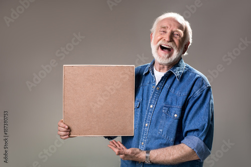 Waist up portrait of happy aging man holding wooden frame, his face expressing unbridled joy. Isolated on grey background