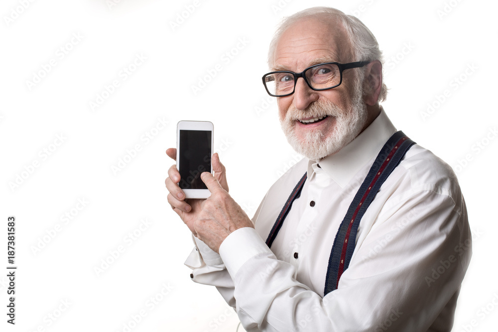Waist up portrait of contented senior pointing his finger at mobile phone and smiling. Copy space in left side. Isolated on background Waist up portrait of contented senior pointing his finger at mobile phone and smiling. Copy space in left side. Isolated on background