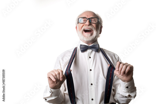 Waist up portrait of joyful old male pulling away his suspenders and laughing. Isolated on background