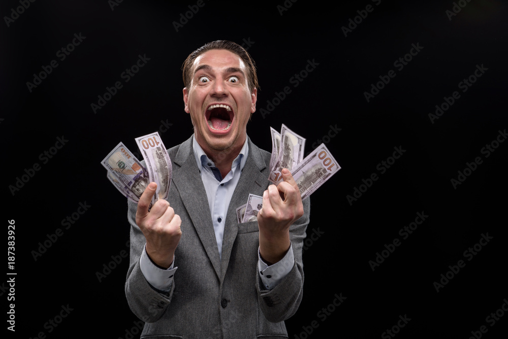 I am crazy about money. Portrait of young insane businessman is holding cash and looking at camera with wide-eyed. He is standing against dark background and screaming. Isolated I am crazy about money. Portrait of young insane businessman is holding cash and looking at camera with wide-eyed. He is standing against dark background and screaming. Isolated