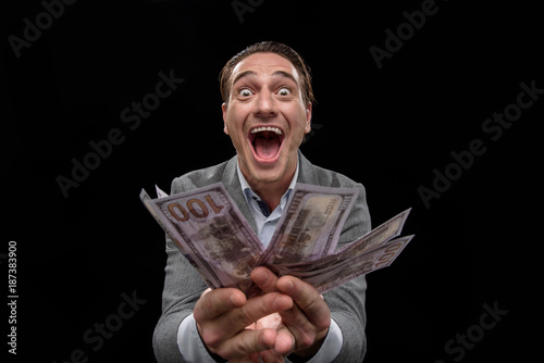 Feeling bid passion about money. Portrait of young crazy entrepreneur is standing and demonstrating dollars while looking at camera with wide-eyed and open mouth. Isolated background