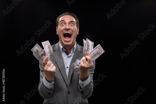 I am crazy about money. Portrait of young insane businessman is holding cash and looking at camera with wide-eyed. He is standing against dark background and screaming. Isolated