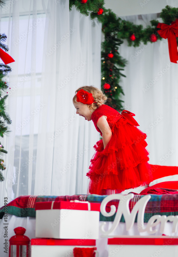 Obraz premium Pretty little girl in red dress looks funny posing in cosy room decorated for winter holidays