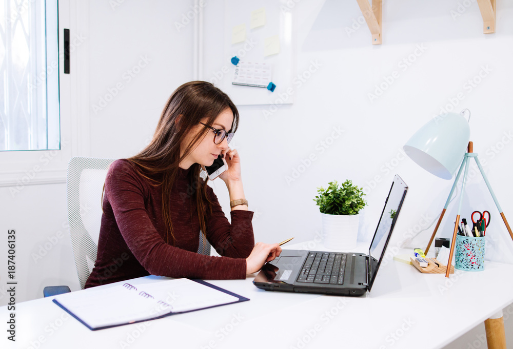 Businesswoman talking on phone while using laptop in office Businesswoman talking on phone while using laptop in office