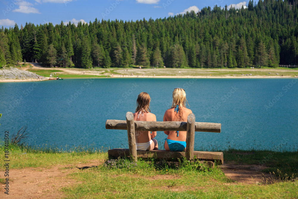 back view of two girls sitting on a bench near a mountain lake and pine trees
