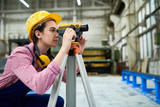 Side view portrait of young female geodesist wearing hardhat looking into optical level mounted on tripod at construction site, copy space