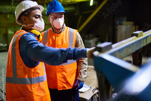 Handsome mixed race technician wearing reflective vest pointing at something while discussing results of joint work with his colleague, interior of spacious production department on background