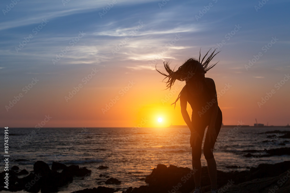Silhouette of flexible girl with dreadlocks on ocean coast during amazing sunset.