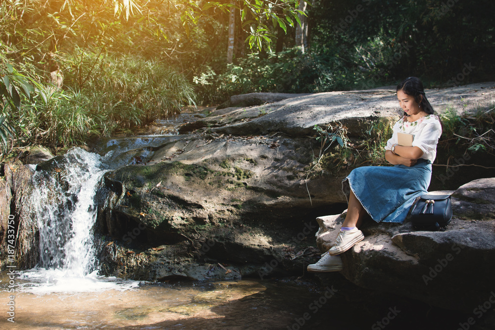 Asian women reading a book sitting on the rock near waterfall in forest background , Relax time and hobby on holiday , soft focus,tone of hipster style Asian women reading a book sitting on the rock near waterfall in forest background , Relax time and hobby on holiday , soft focus,tone of hipster style